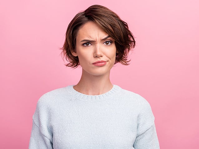 A person with short brown hair wearing a light gray sweater stands against a pink background, displaying a quizzical expression with one eyebrow raised.