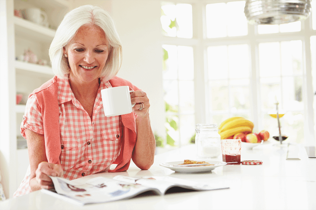 Older woman sitting at a table sipping from a mug while reading a magazine and smiling